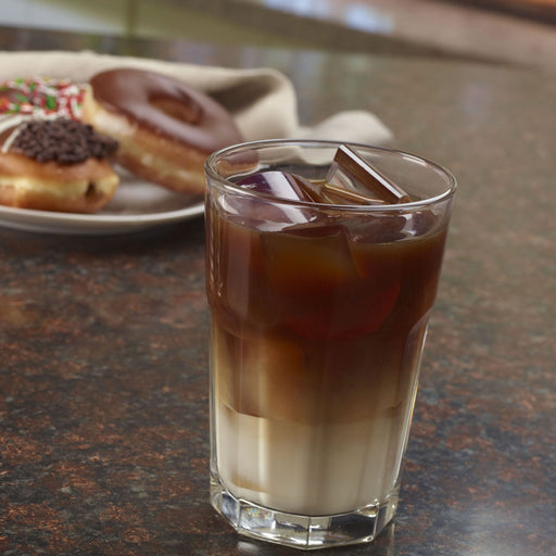 Ice-filled glass of coffee on a table with donuts in the background.