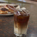 Ice-filled glass of coffee on a table with donuts in the background.